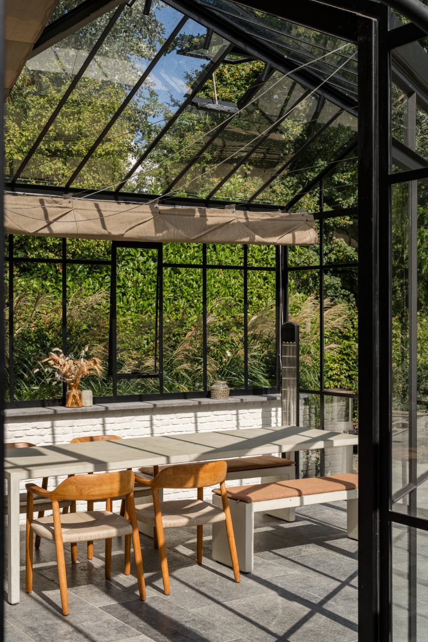 Houten eettafel en stoelen onder het glazen dak van een stalen orangerie met zicht op het groen