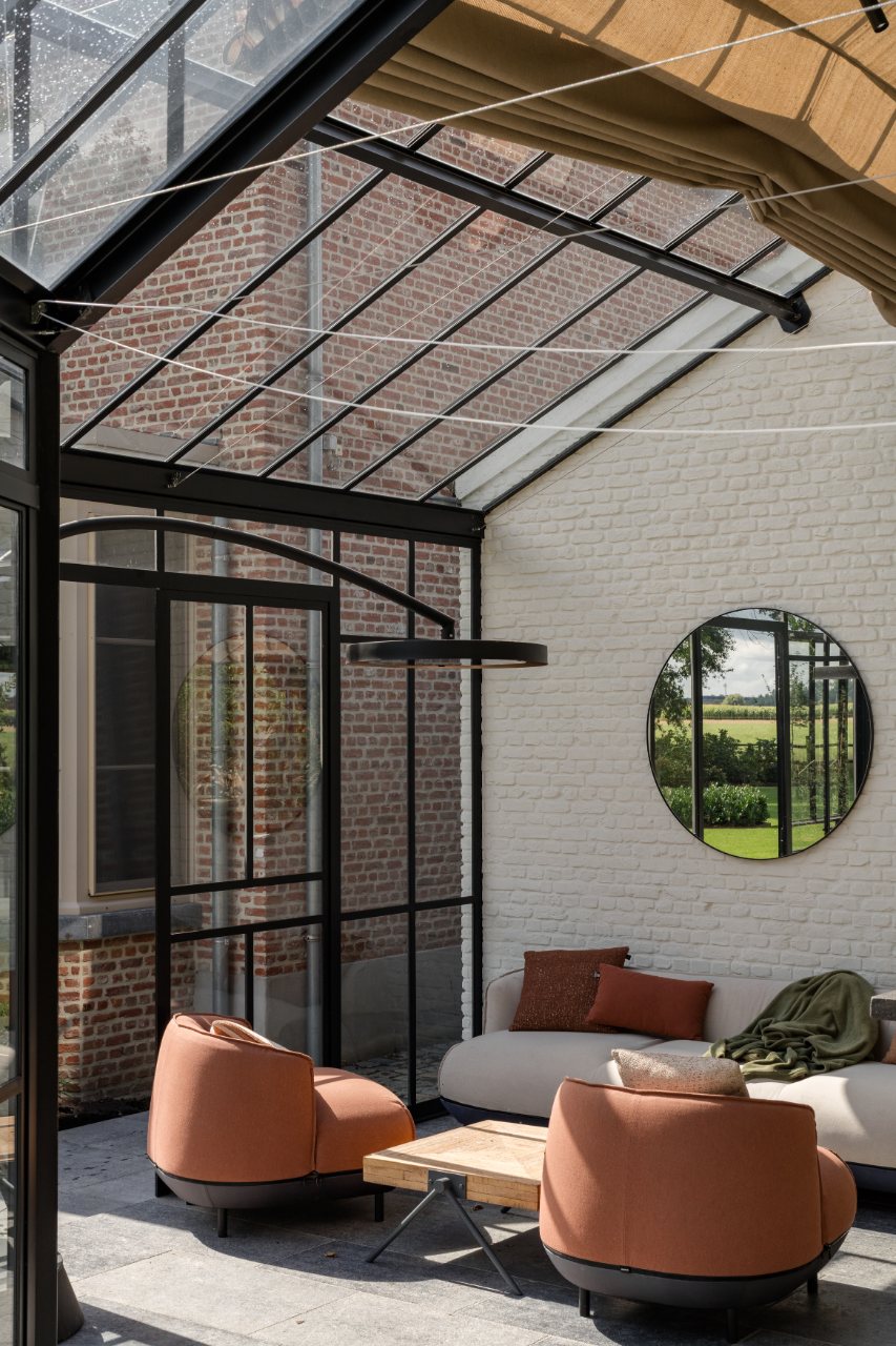 Interieur van een stalen orangerie met glazen dak, loungezetels en een rond raam