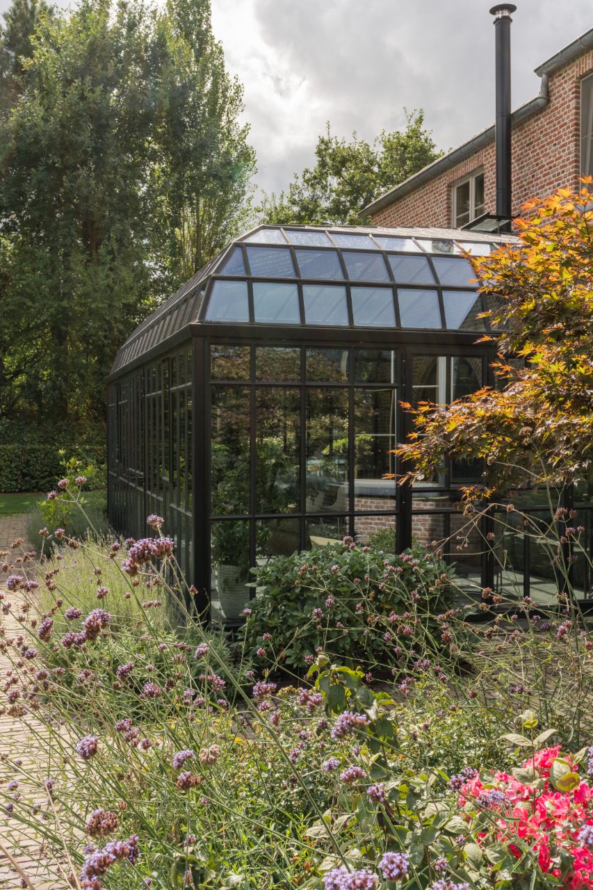 Zwarte stalen orangerie met glazen dak aan een woning, omringd door een bloemrijke tuin