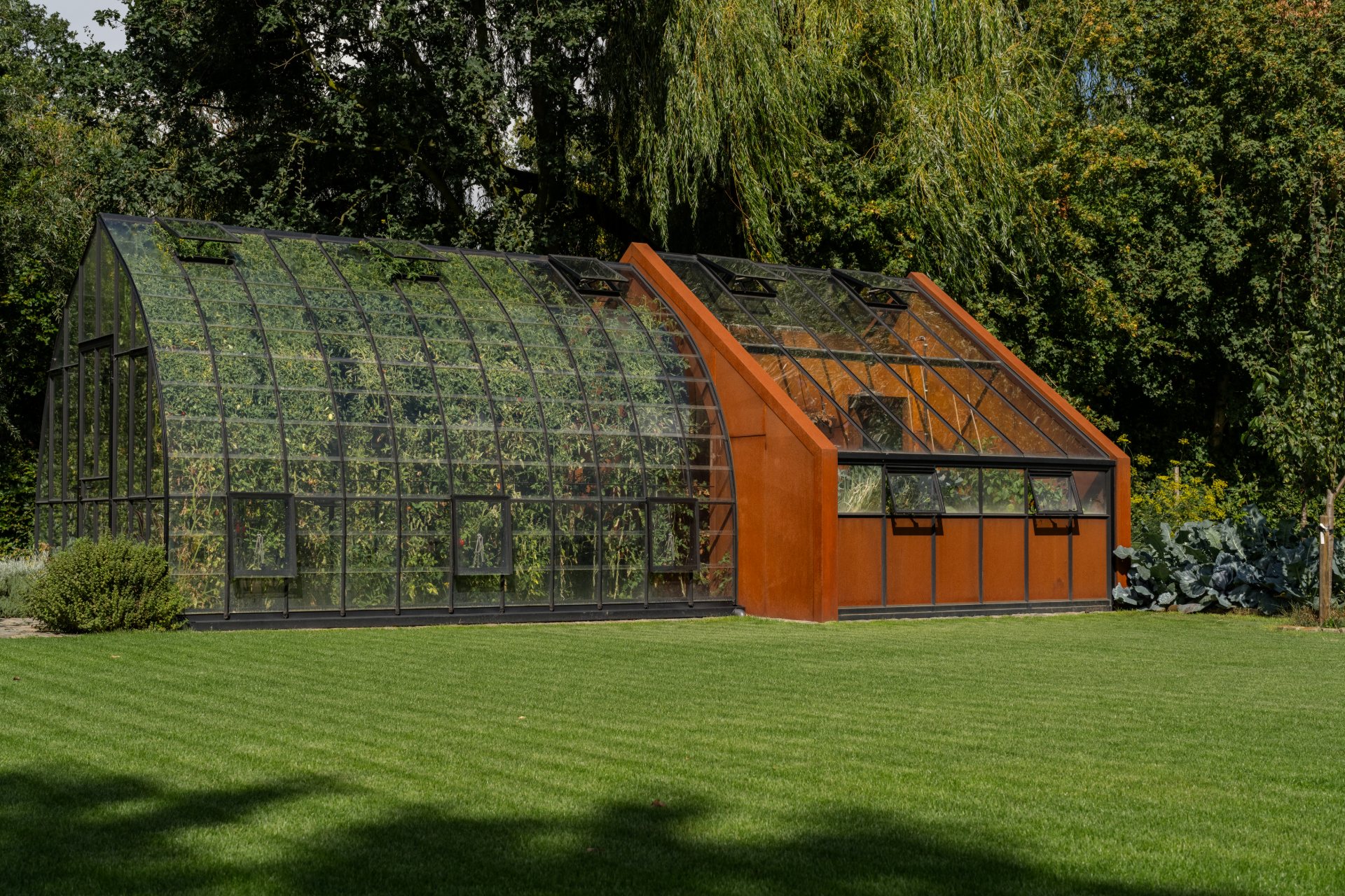 Tuinserre in zwart staal en glas met een cortenstalen berging in een groene tuin